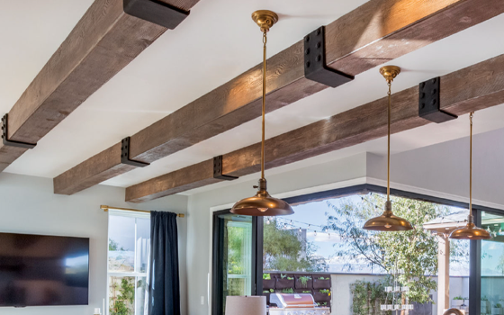 Large wooden beams with black straps sit on a white ceiling. Gold light fixtures hang down over brightly lit living room with window in background.